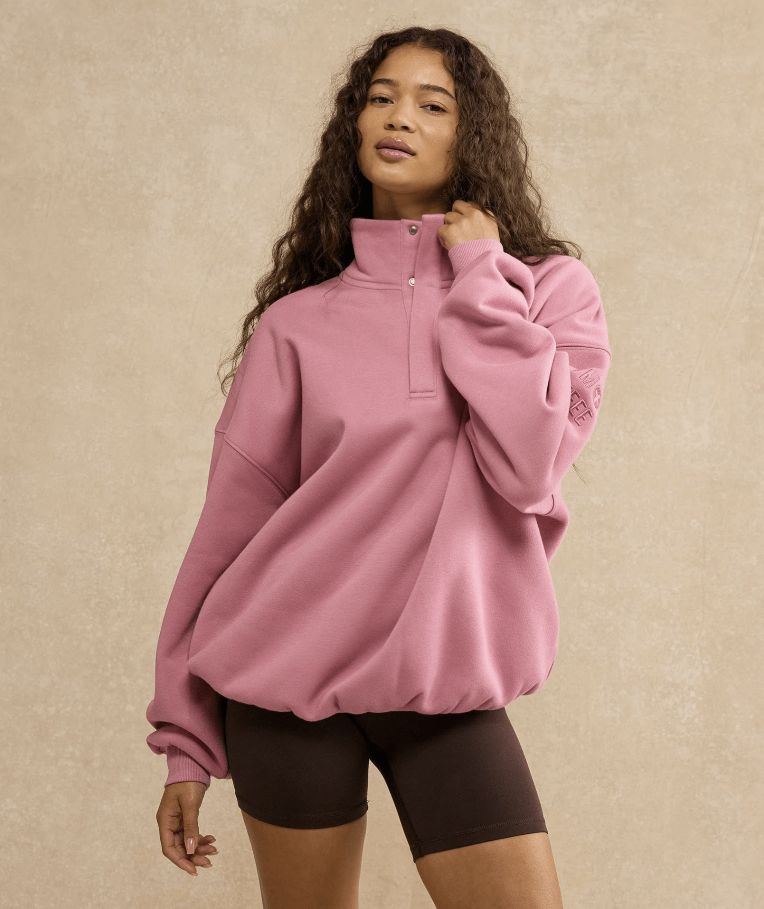 Woman wearing a loose pink oversized sweatshirt with a high collar and brown bike shorts, posed against a beige textured backdrop.