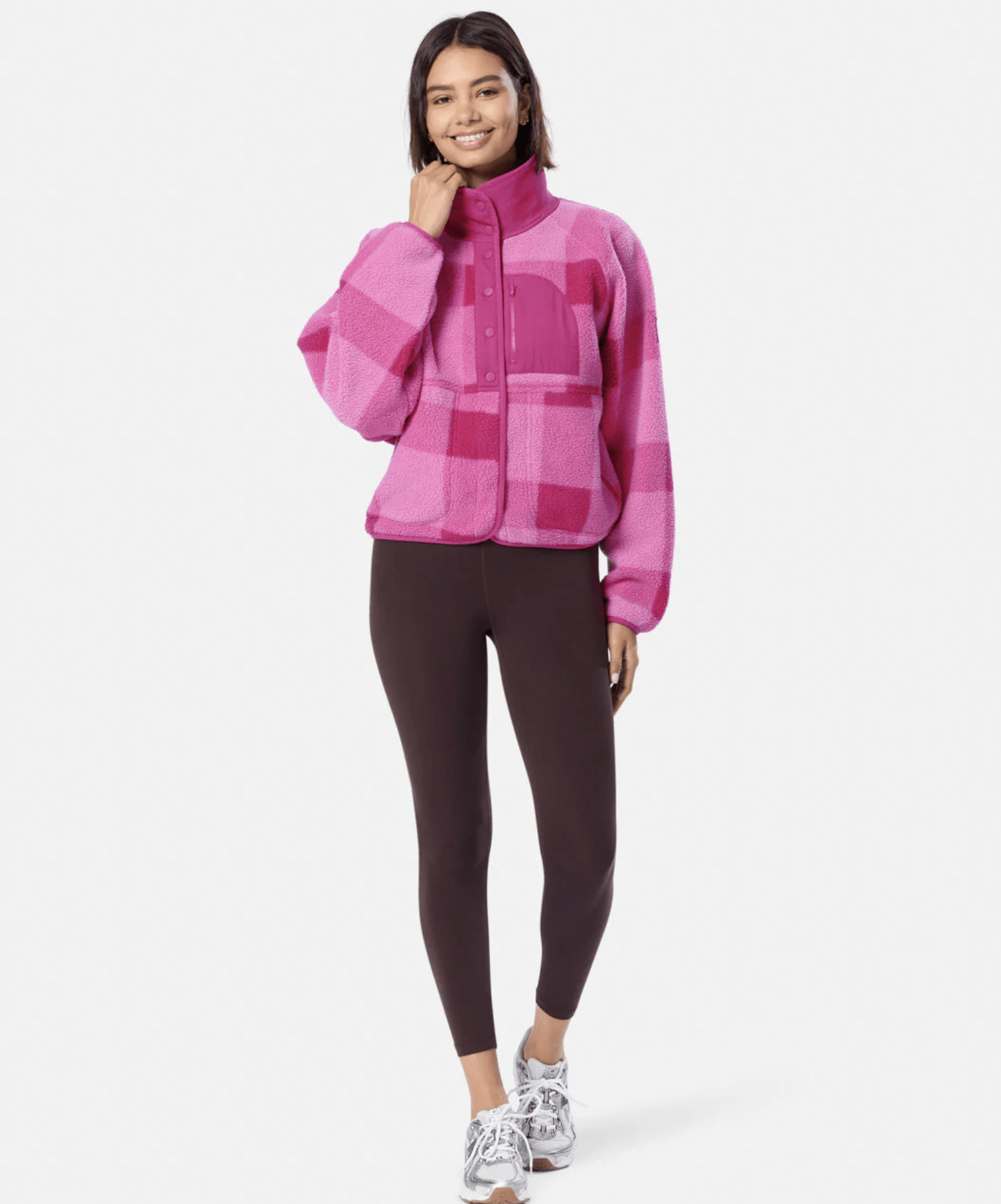 Woman wearing a bright pink plaid fleece jacket with a high collar, front snap buttons, and a chest zip pocket; paired with dark leggings and gray sneakers.