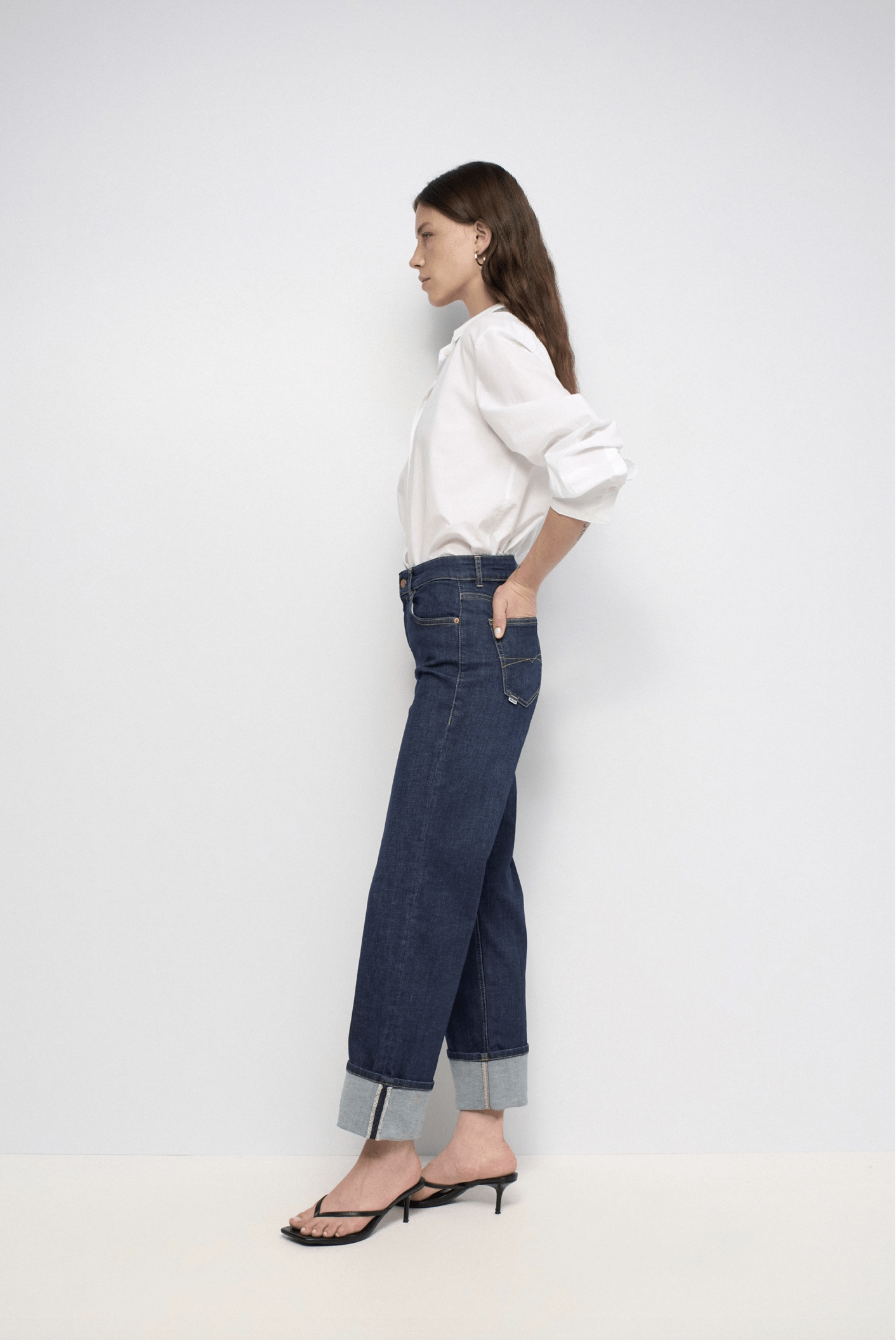 Woman standing in profile facing left, wearing a white button-down shirt, rolled-cuff blue jeans, and black sandals.